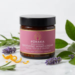 Pūraka Brightening Soothing Hand Cream amber glass jar surrounded by fresh lavender, orange peel and green leaves on a marble surface